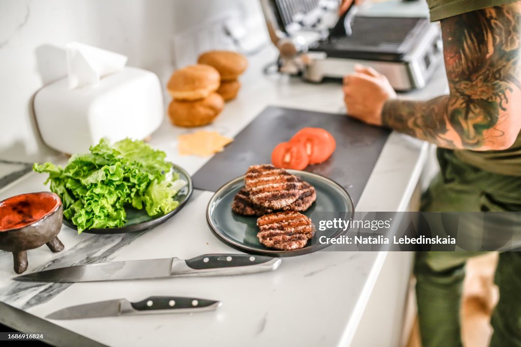 Homemade burgers: Minced meat and salad sizzle on the electric grill, with tomatoes on the kitchen table.