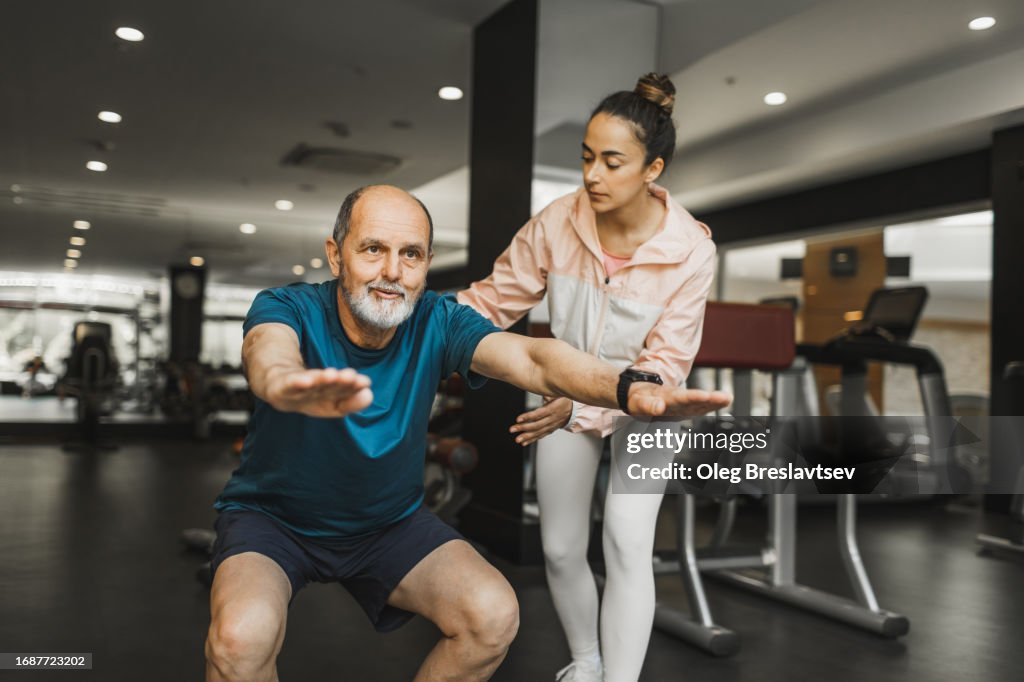 Senior man doing squats in gym for body and muscles rehabilitation and young coach assisting him