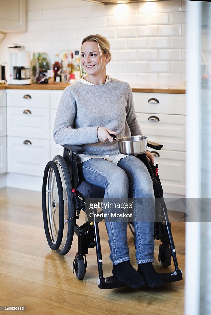 Happy disabled woman in wheelchair looking away while holding saucepan at kitchen