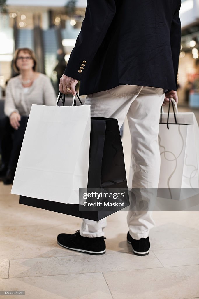 Low section of man carrying shopping bags with woman in background at mall