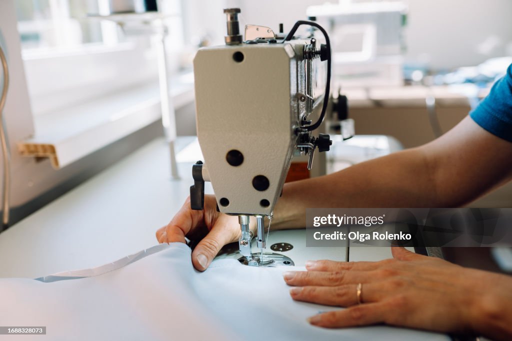 Artful tailoring in atelier: unrecognizable mature woman hands with ring work on sewing machine with white textile on blurred background