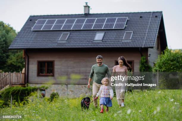 young family with dog on a walk through the meadow near their family home with solar panels on the roof. - family technology stock-fotos und bilder