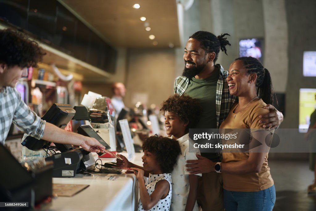 Happy black family buying movie tickets in cinema.