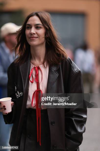 Alexa Chung is seen wearing a knee-length black leather jacket with silver gemstone details, underneath a rose top with red ribbons and a black...