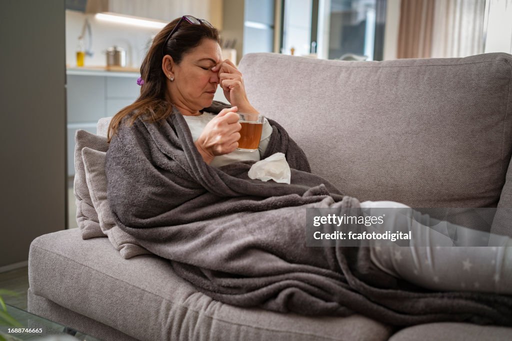 Sick woman resting on sofa drinking hot tea and massaging her nose