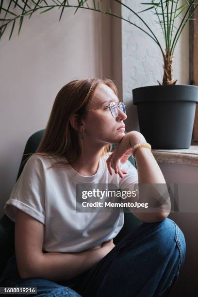 upset depressed student girl looking out window, feeling sad, coping with breakup. unhappy redhead woman thinking about life, looking into distance, feeling lonely at home - procrastination stock-fotos und bilder