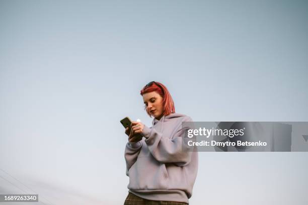happy young woman using smart phone standing outdoors against blue sky - guay fotografías e imágenes de stock