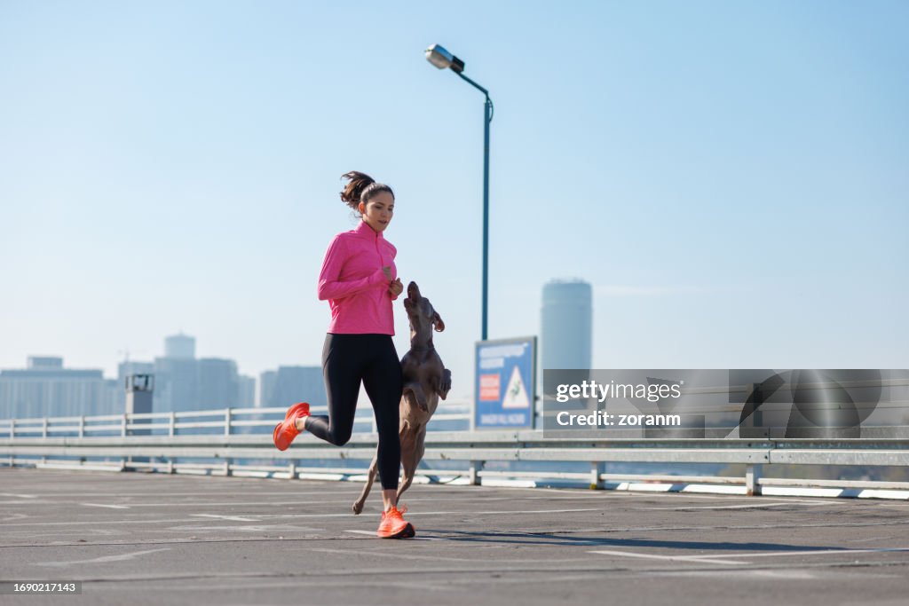 Young woman running with her dog jumping next to her on empty parking lot in the city