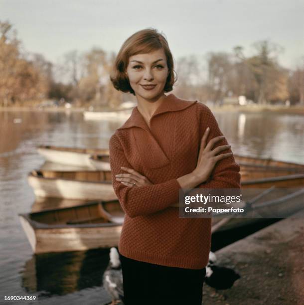 Posed portrait of a female fashion model wearing a red cable knit jumper with open collar, she stands beside a boating lake in England, 17th...