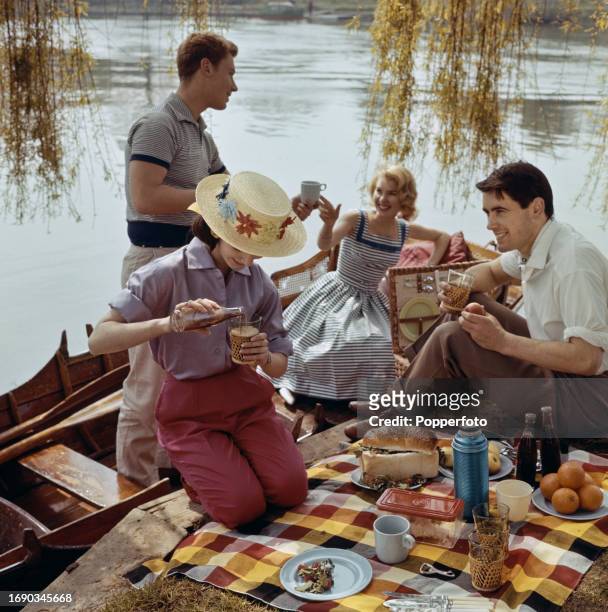 Two young women and their male partners enjoy a picnic lunch of salad, bread, fruit and bottles of Pepsi-Cola beside their rowing boat moored to the...