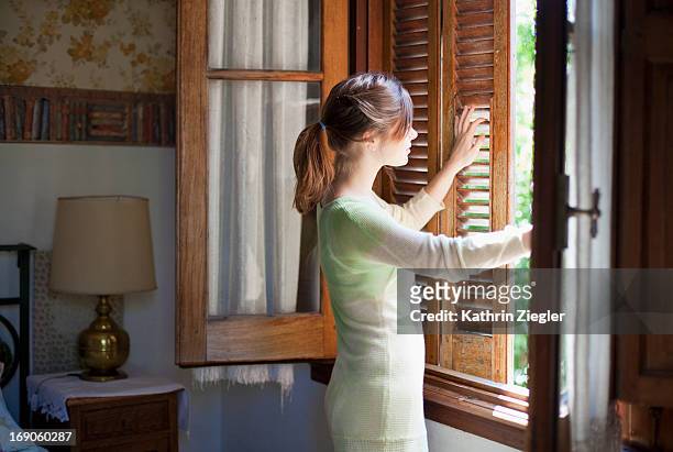young woman unshuttering the window - fensterladen stock-fotos und bilder