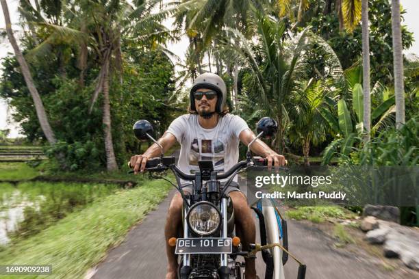 multiracial male surfer riding motorcycle along small road in bali - indonesia surfing stock pictures, royalty-free photos & images