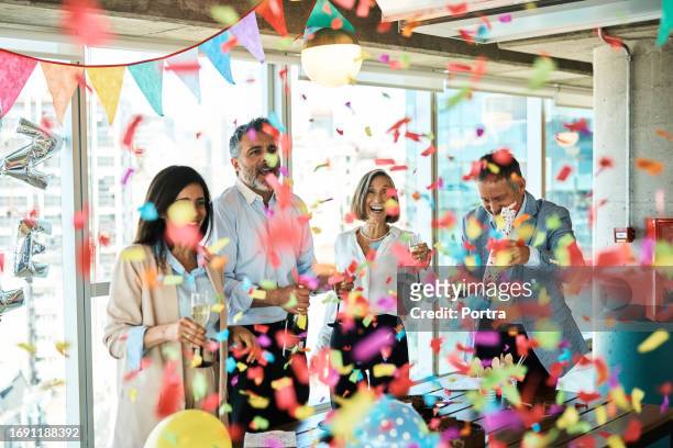 mature business people celebrating with confetti party popper in office - birthday office stockfoto's en -beelden