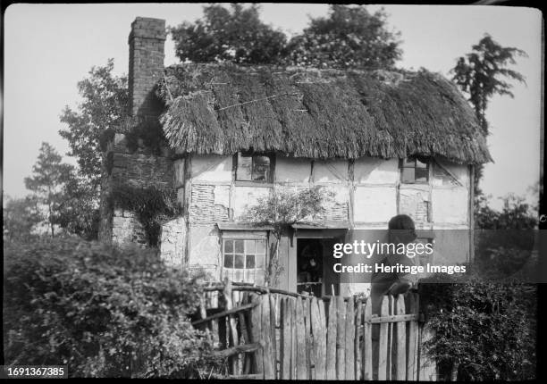 Woolhope, Herefordshire, 1930-1949. The front of a timber framed thatched cottage in Woolhope with a woman standing in the garden looking back at the...