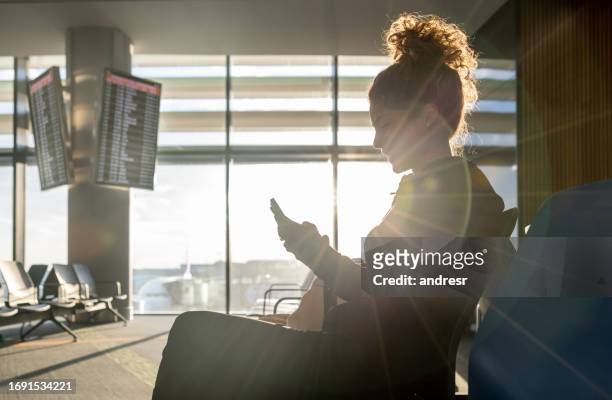 female traveler texting on her cell phone while waiting by the gate at the airport - travel insurance stock pictures, royalty-free photos & images