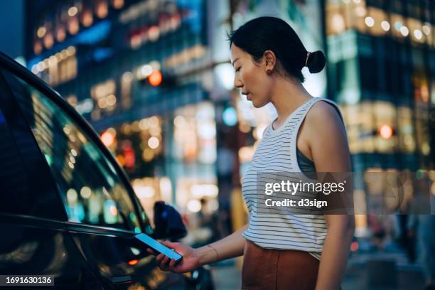 young asian woman using mobile app device on smartphone to unlock the doors of her intelligence car in the city at night, against illuminated buildings. wireless and modern technology. smart car with digital car key concept - car rental stock pictures, royalty-free photos & images