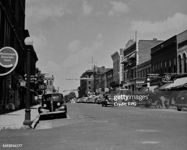 Cars parked along the sidewalk, among the store signs in view are the 'Western Auto Associate Store', 'Russell & Son Market', 'Franklin's Studio' and...