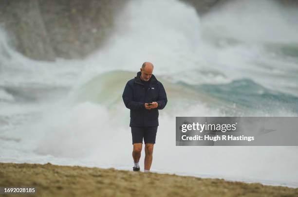 Man studies his mobile phone in front of stormy waves at Gyllyngvase Beach on September 20, 2023 in Falmouth, England. Parts of England are...