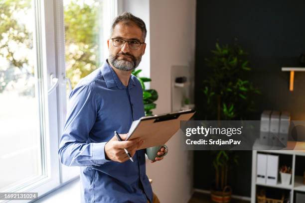 hombre de negocios adulto medio haciendo una lluvia de ideas mientras está parado cerca de la ventana, mientras sostiene el café y el portapapeles - tablilla con sujetapapeles fotografías e imágenes de stock