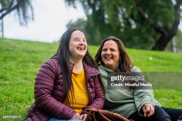 woman with down syndrome laughing while sitting in grass with mother - syndroom van down stockfoto's en -beelden