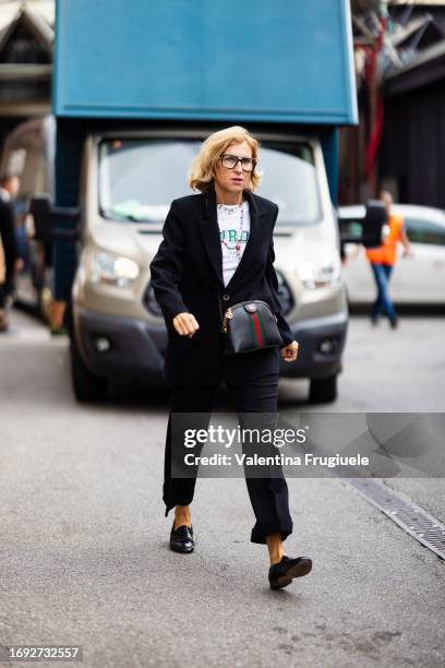 Guest is seen wearing black and gold glasses, a white t-shirt white green littering details, a black blazer, black trousers, black shiny leather...