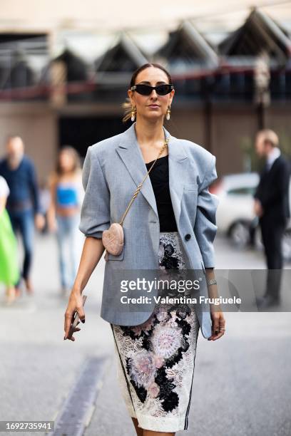 Elisa Taviti is seen wearing black sunglasses, gold and pearls earrings, a black top, a grey oversized blazer, a Dior heart-shaped cross-body pink...