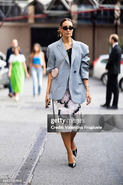 Elisa Taviti is seen wearing black sunglasses, gold and pearls earrings, a black top, a grey oversized blazer, a Dior heart-shaped cross-body pink...