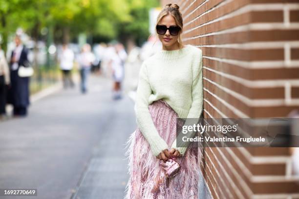 Ginta is seen wearing a mint green sweater, black sunglasses, a pink midi dress embellished with feathers and sequins and a pink shiny mini bag with...