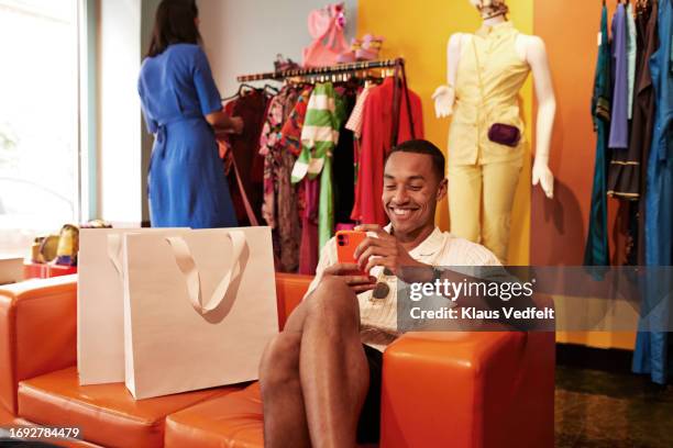 happy young man using mobile phone on sofa in store - shopping center - fotografias e filmes do acervo