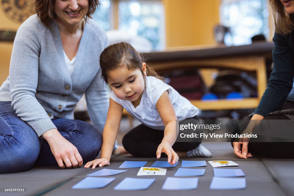 Niña en edad preescolar juega un juego de cartas con la madre y el terapeuta ocupacional