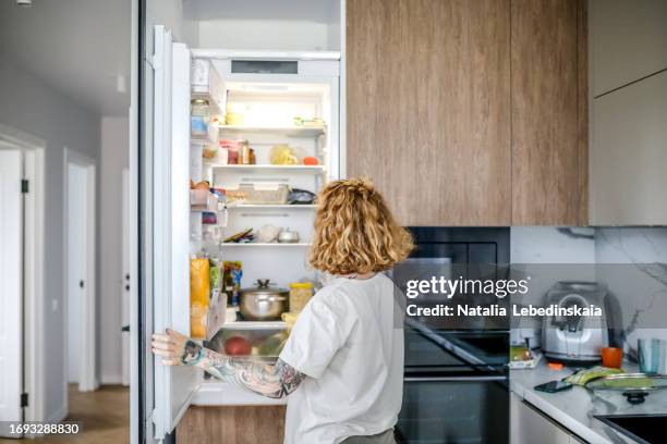 red-haired woman in front of open refrigerator at home - platzieren stock-fotos und bilder