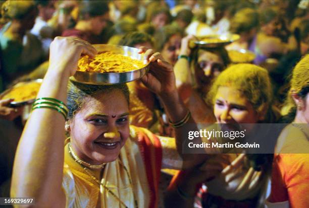 Women in white saris throwing powdered yellow turmeric and coconut chunks over revellers at a Hindu festival from silver platters on their...