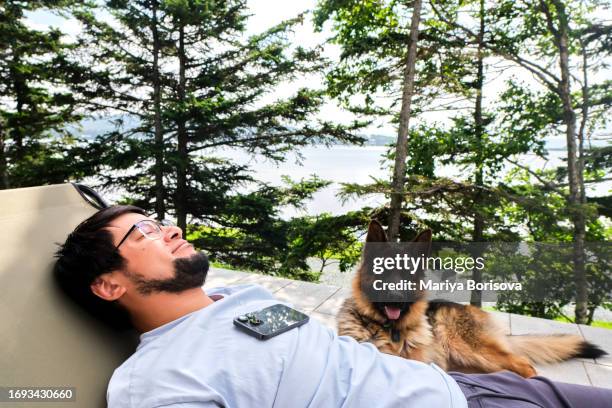 a man lies on a sun lounger with a phone on the veranda of his own house. a dog sits nearby. - schnarchen mann stock-fotos und bilder