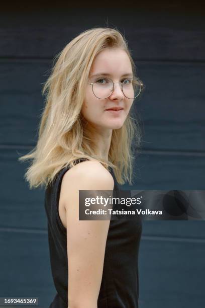 portrait of teenage girl with fair hair and eyeglasses in black tank top standing in front of dark gray wall - regarder par dessus son épaule photos et images de collection
