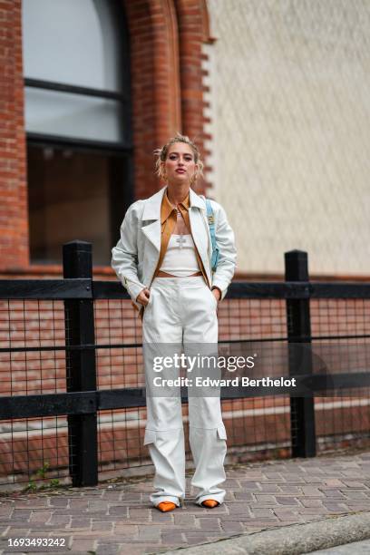 Emily Sindlev wears earrings, a leather jacket, a brown leather shirt, a white low-neck top, pale blue denim jeans, orange pointed shoes, outside...