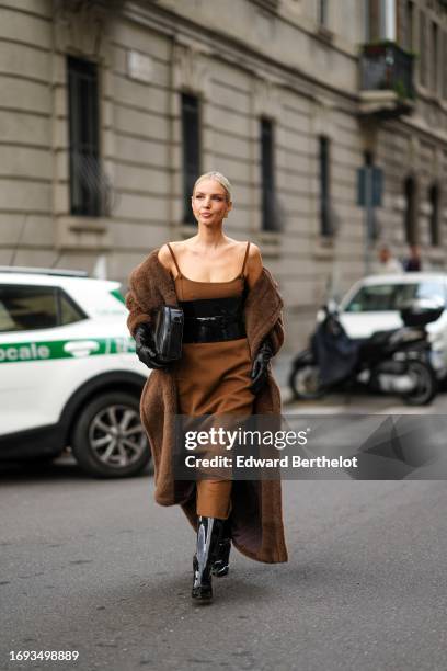Leonie Hanne wears a brown long dress, a black leather shiny corset, black leather gloves, a brown long fluffy winter coat, black pointed boots, a...