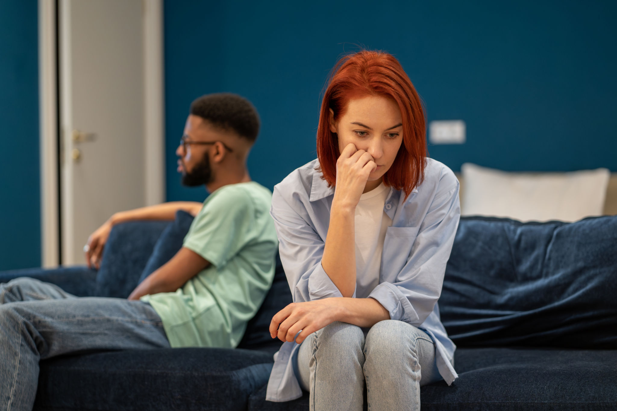 Upset diverse couple wife and husband sit separately on sofa at home not talking after quarrel Upset diverse couple wife and husband sit separately on sofa at home not talking after quarrel