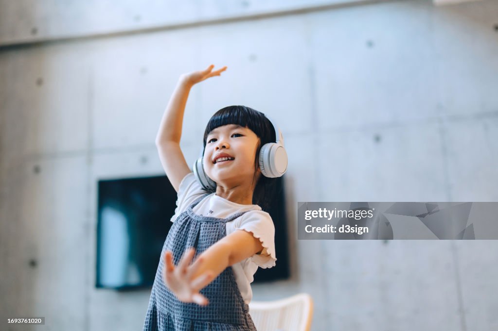 Happy little Asian girl listening to music through headphones while dancing. Having fun singing and dancing in the living room at home