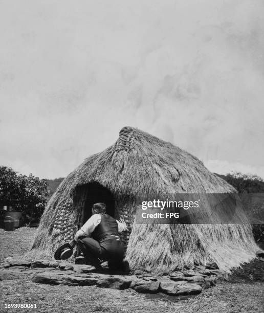 Man, wearing a black waistcoat over a white shirt and holding a black hat, crouches at the entrance to a hale, a traditional grass hut in Hawaii,...