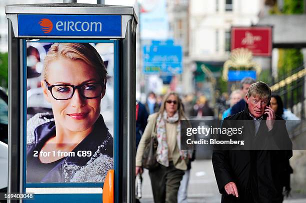 Pedestrian speaks on a mobile phone as he passes an advert for Specsavers Optical Group Ltd. On the door of a fixed-line public telephone booth,...