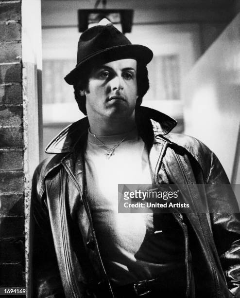 American actor Sylvester Stallone leans against a doorway in a hat and a leather jacket in a still from the film, 'Rocky,' directed by John G....