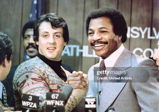 American actors Sylvester Stallone and Carl Weathers grip hands and smile together during a press conference in a still from the film, 'Rocky,'...