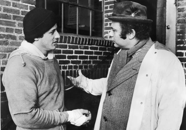 American actors Sylvester Stallone , wearing training clothes, and Burt Young talk outdoors in a still from the film, 'Rocky,' directed by John G....