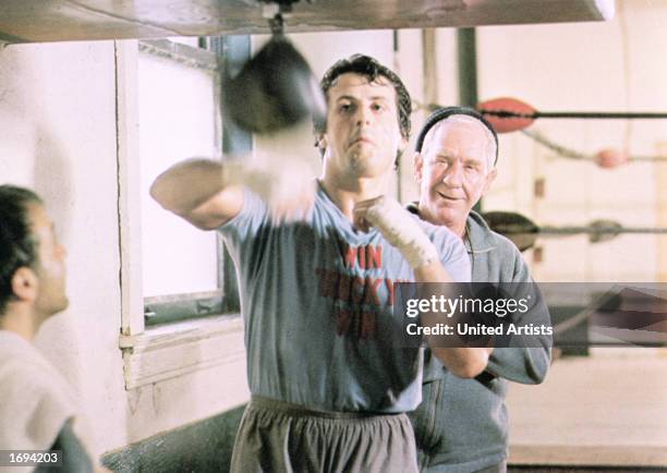 American actor Sylvester Stallone strikes at a punching bag in a boxing ring while his coach, Burgess Meredith , watches during training at a...