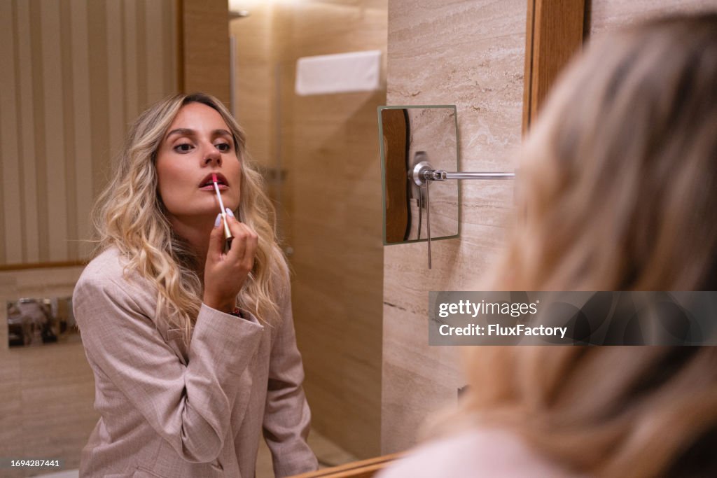 Mujer caucásica aplicando brillo labial, mientras se observa en el espejo del baño