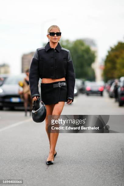 Caroline Daur wears sunglasses, a black cropped jacket, a mini skirt with a leather belt, a moon shaped Prada bag, pointed shoes, outside Prada,...