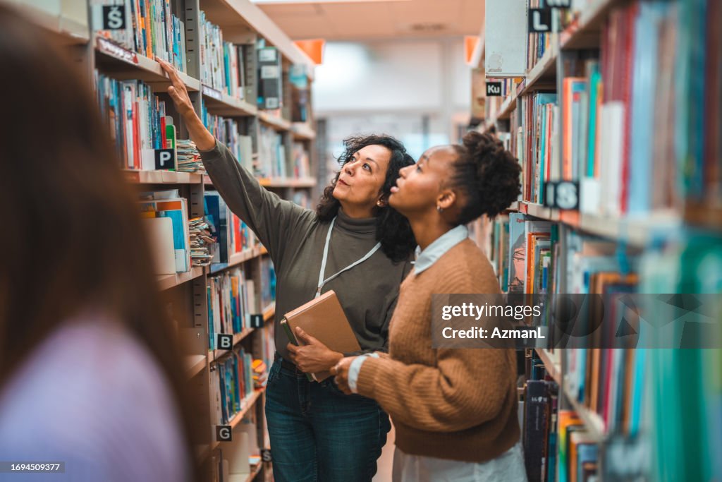 Hispanic Student Searching For A Book With A Librarian