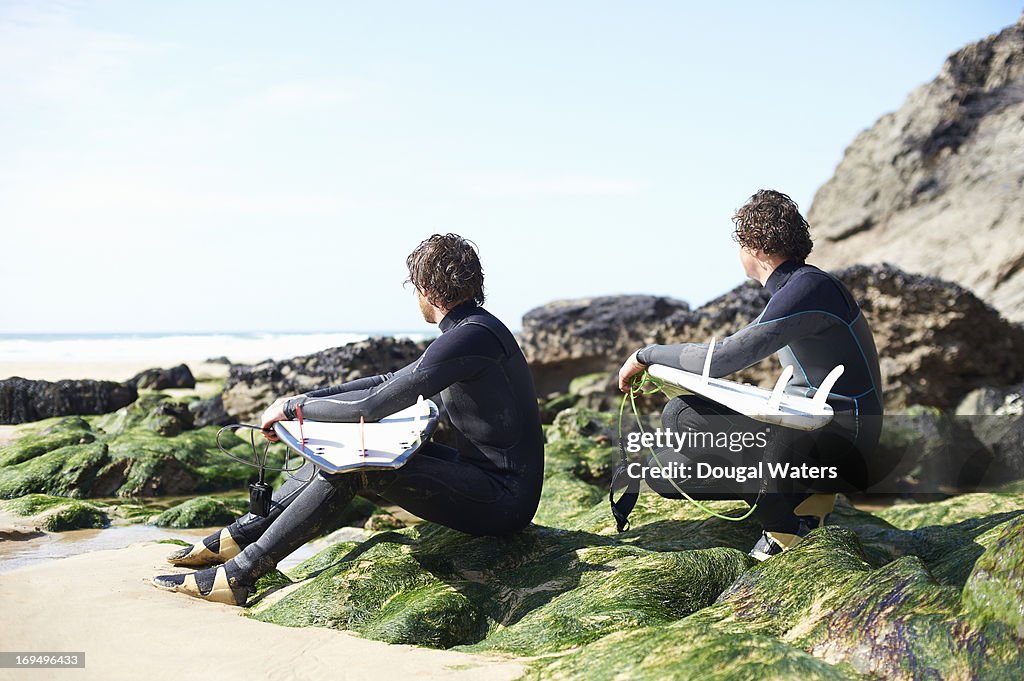 Surfers sitting on rocks beside sea.