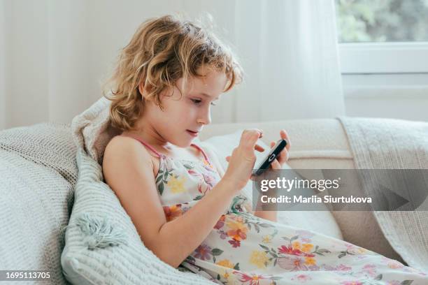 cute little girl using digital tablet technology device lying on sofa alone. small child hold mobile phone surfing internet play game at home. children tech addiction concept. - addiction mobile and laptop stockfoto's en -beelden