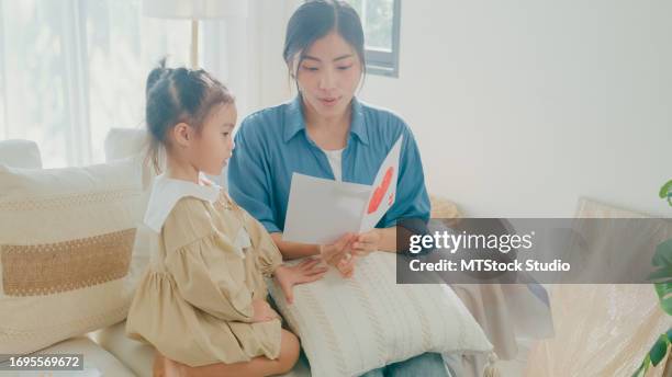 young asian family mom and daughter hugging and reading handmade greeting card with heart having fun on sofa in living room during holiday celebration mothers day at home. family happy moment. - receiving paper stock pictures, royalty-free photos & images